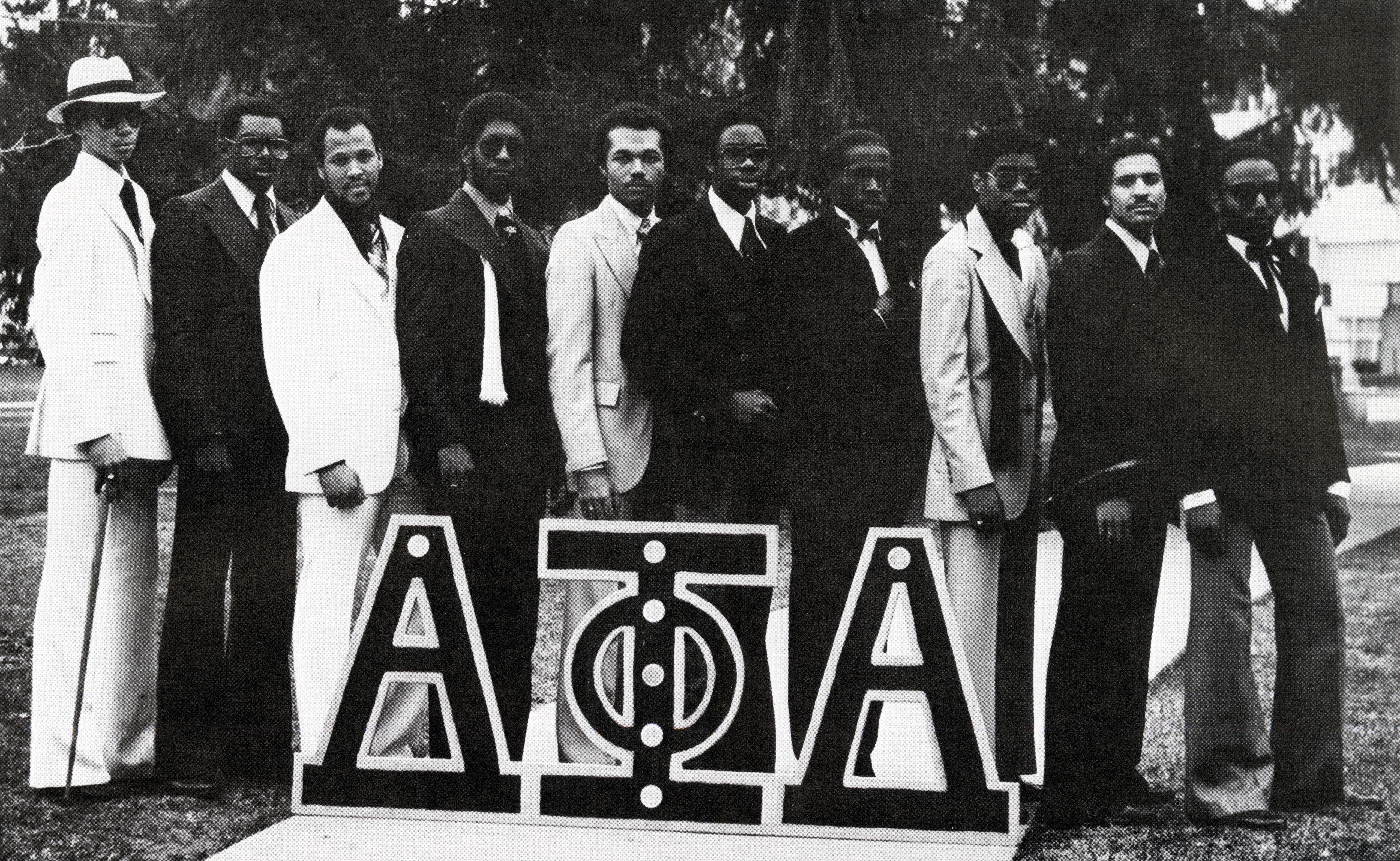 Black and white group photo of the brothers of the Alpha Phi Alpha fraternity at Dartmouth in 1979. All men pictured are wearing suits and standing behind a sign on the ground showing the letters of the fraternity.