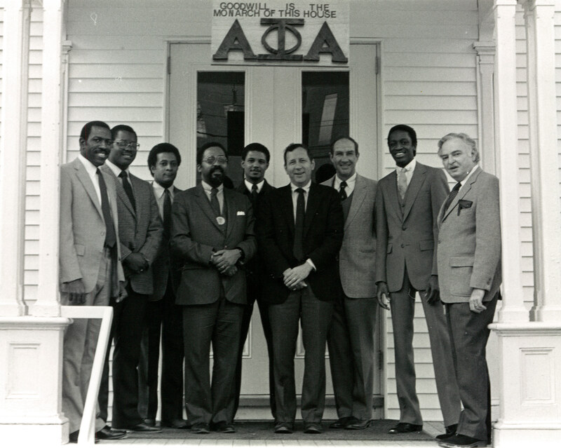 Black and white photo of nine men, including Alpha Phi Alpha brothers and Dartmouth President John G. Kemeny, standing on the porch of the Alpha Phi Alpha fraternity house. Above the house front door are the fraternity letters and a sign that reads "Goodwill is the monarch of this house."