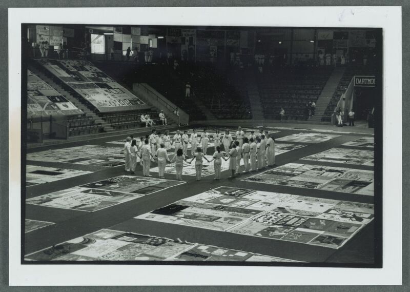 A panoramic view of Thompson Arena, with a group of people dressed in white encircling a section of quilt panels and holding hands.