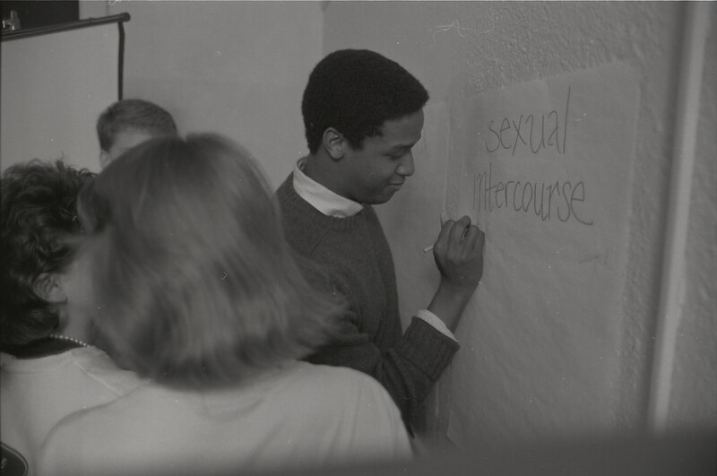 A smiling student writes "sexual intercourse" in large letters on a poster.