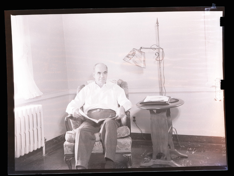 Ernest Martin Hopkins, a balding, white man with thick eyebrows and a severe expression, sits on an armchair in a brightly lit room, holding an open magazine in his lap.