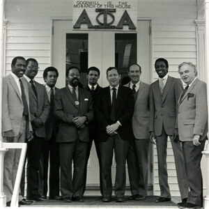 Black and white photo of nine men, including Alpha Phi Alpha brothers and Dartmouth President John G. Kemeny, standing on the porch of the Alpha Phi Alpha fraternity house. Above the house front door are the fraternity letters and a sign that reads "Goodwill is the monarch of this house."